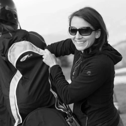 Frau mit Sonnenbrille bereitet Ausrüstung für Paragleiten vor, lächelnd, Outdoor mit Bergblick bei Austriafly Flugschule
