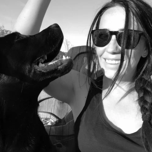 Frau mit Sonnenbrille und Zopf lächelt neben schwarzem Hund im Freien vor Bergkulisse bei Sonnenschein