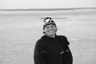 Junger Mann mit Mütze und Outdoorjacke lächelt vor einer weiten, offenen Landschaft in Schwarz-Weiß Aufnahme.