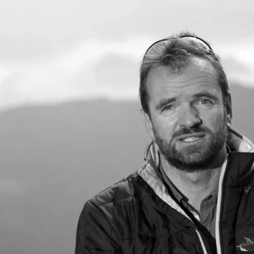 Porträt eines Mannes mit Bart und Jacke mit Austriafly-Logo vor Bergkulisse in Schwarz-Weiß