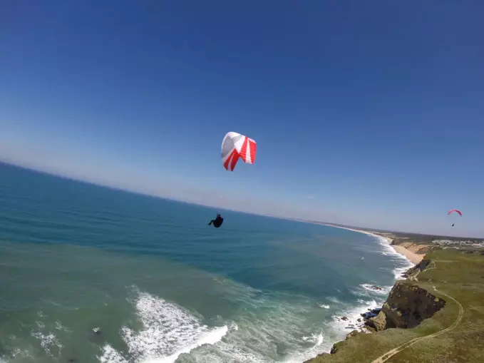 Paragleiter mit rotem und weißem Schirm fliegt über Küste mit Klippen und blauem Meer bei klarem Himmel