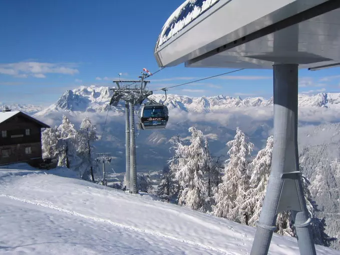 Winterliche Berglandschaft mit Gondelbahn, schneebedeckten Bäumen und Bergen im Hintergrund bei klarem Himmel.