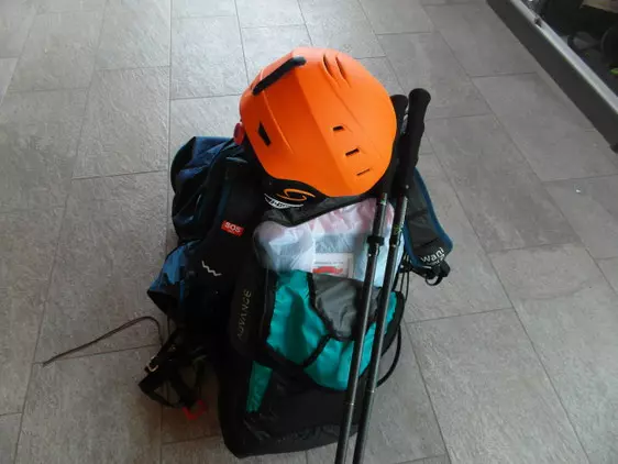 Rucksack mit orangefarbenem Helm und Wanderstöcken auf grauem Fliesenboden, Ausrüstung für Gleitschirmfliegen oder Wandern