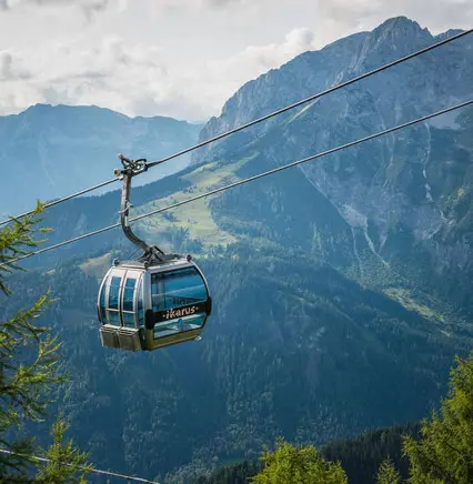 Gondelbahn mit der Aufschrift ikarus vor Bergkulisse in den Alpen bei Wörgl, Österreich, umgeben von Nadelbäumen.